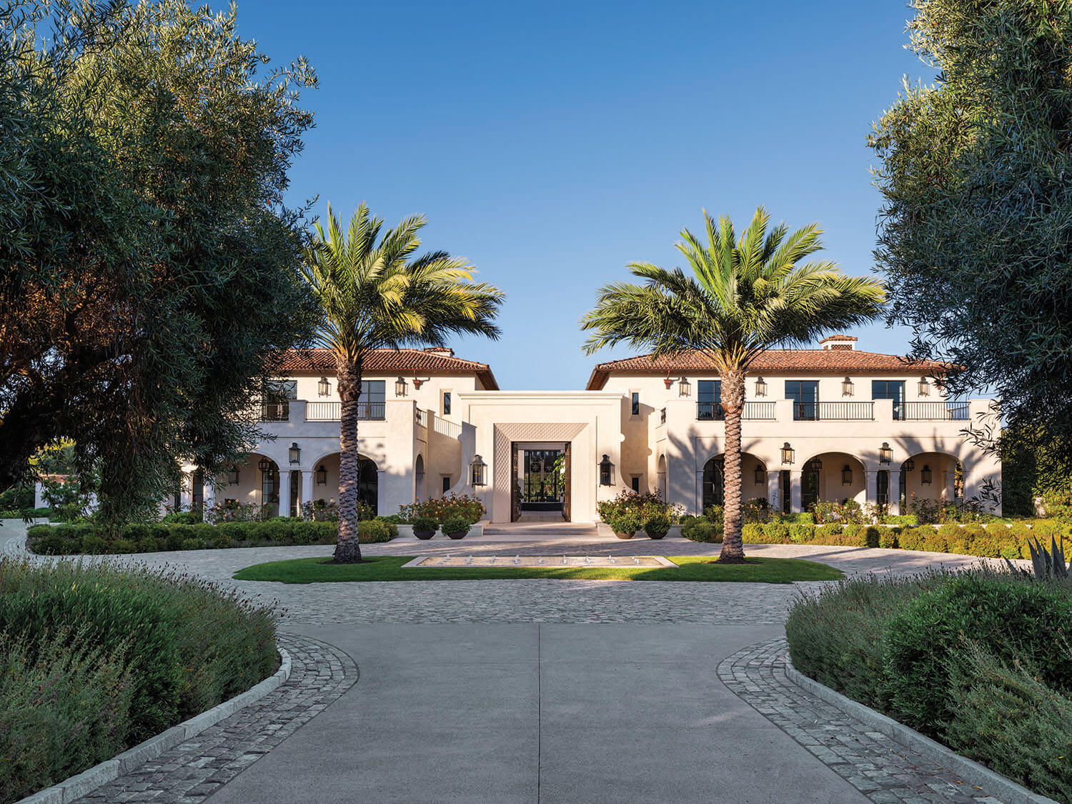 Large Mediterranean-style mansion with white exterior, red-tiled roof, arched entryway, and palm trees flanking a circular driveway under a clear blue sky.
