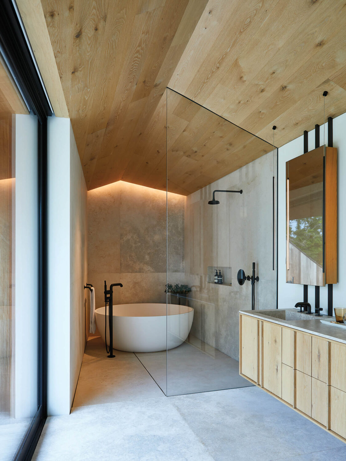 Modern bathroom with wood ceiling, freestanding bathtub, glass shower divider, and light wood vanity under a large mirror. Natural light enters through a floor-to-ceiling window on the left.