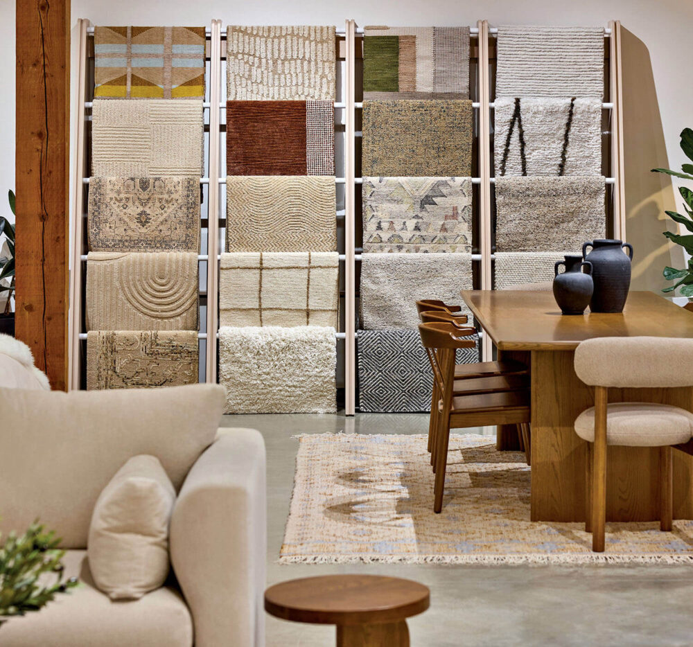 Display of various area rugs on shelves behind a wooden dining table with chairs and a beige sofa in a modern showroom setting.