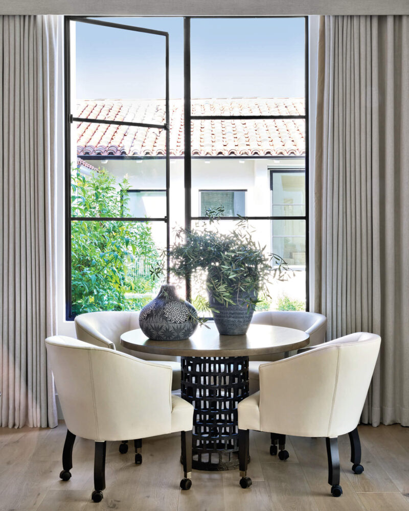 Round table with decorative vases, surrounded by four white chairs, placed in front of a large window with open panes and closed curtains, overlooking a tiled roof and greenery outside.