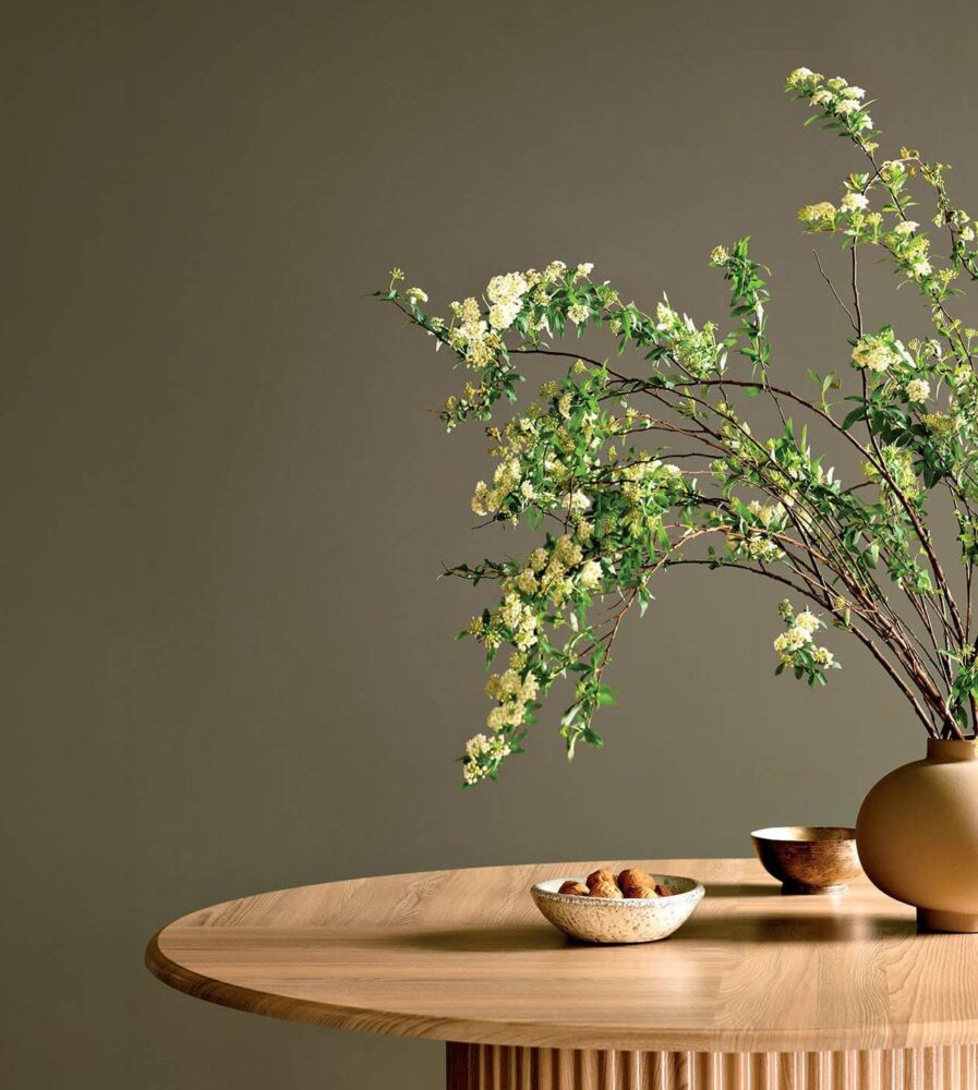 A wooden table with a vase of flowering branches and two small bowls against a plain beige wall.