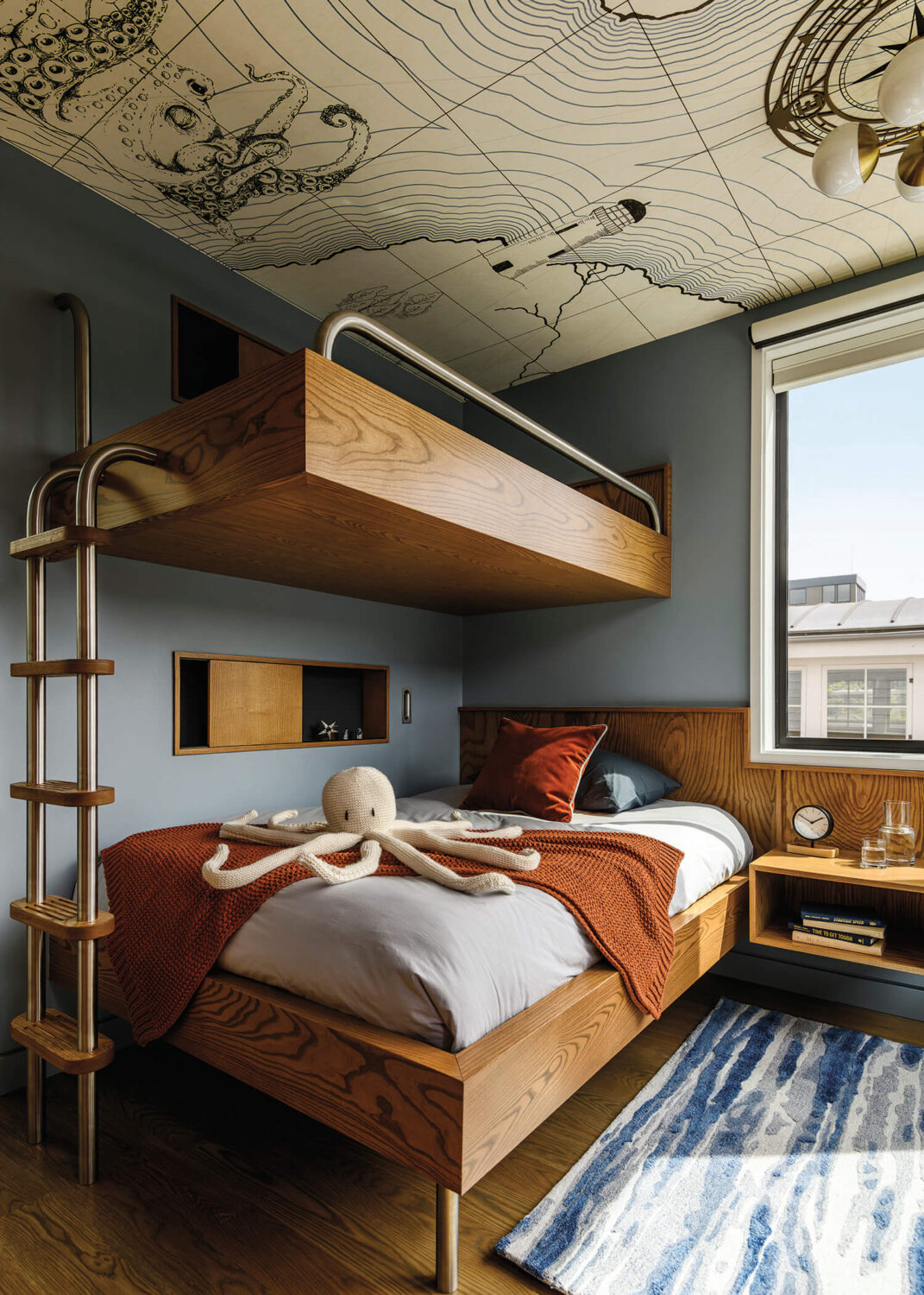 Modern kids’ bedroom with wooden bunk beds, a stuffed octopus toy on the lower bed, blue walls, and a ceiling featuring a nautical map illustration. Natural light comes through a window.