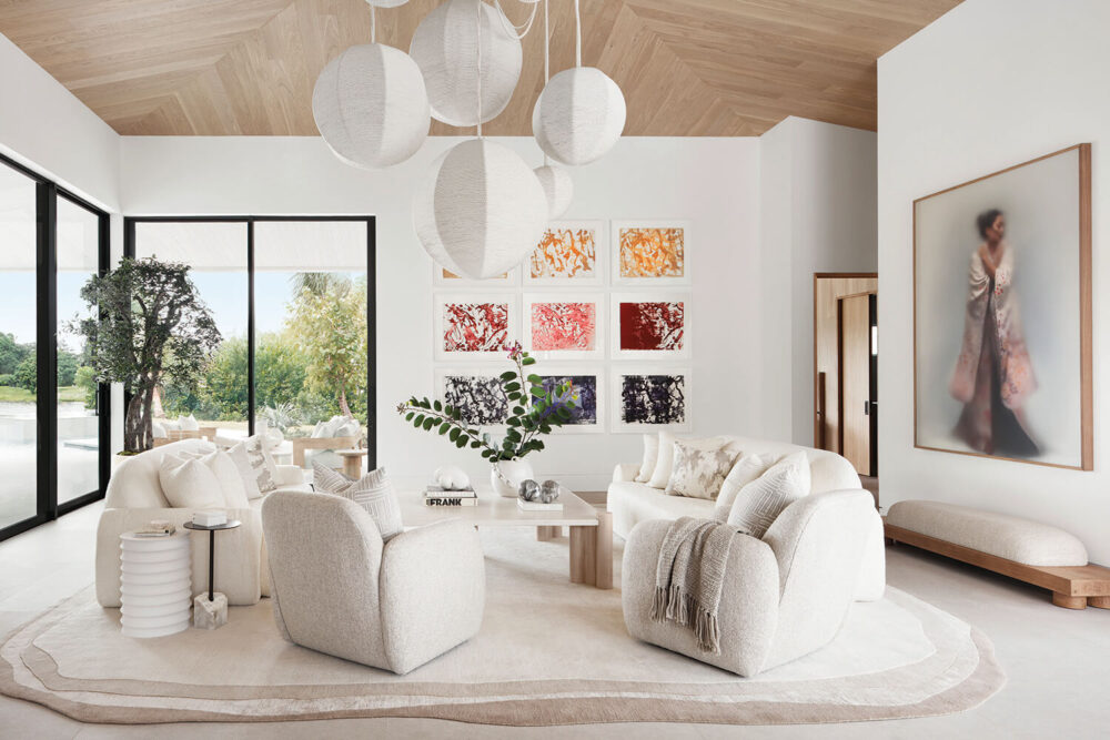Modern living room with white and beige furniture, abstract art, large globe light fixtures, glass doors to the outside, and a blurred portrait on the wall.