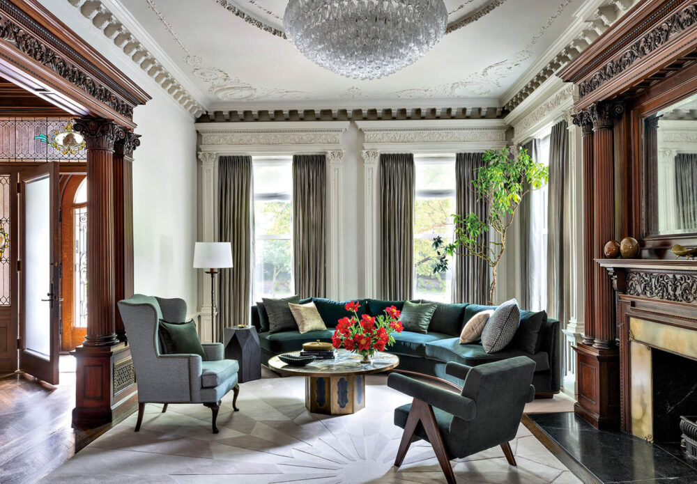 Elegant living room with ornate ceiling, large windows, dark wood trim, green sectional sofa, armchairs, fireplace, and a round coffee table with a red flower arrangement.