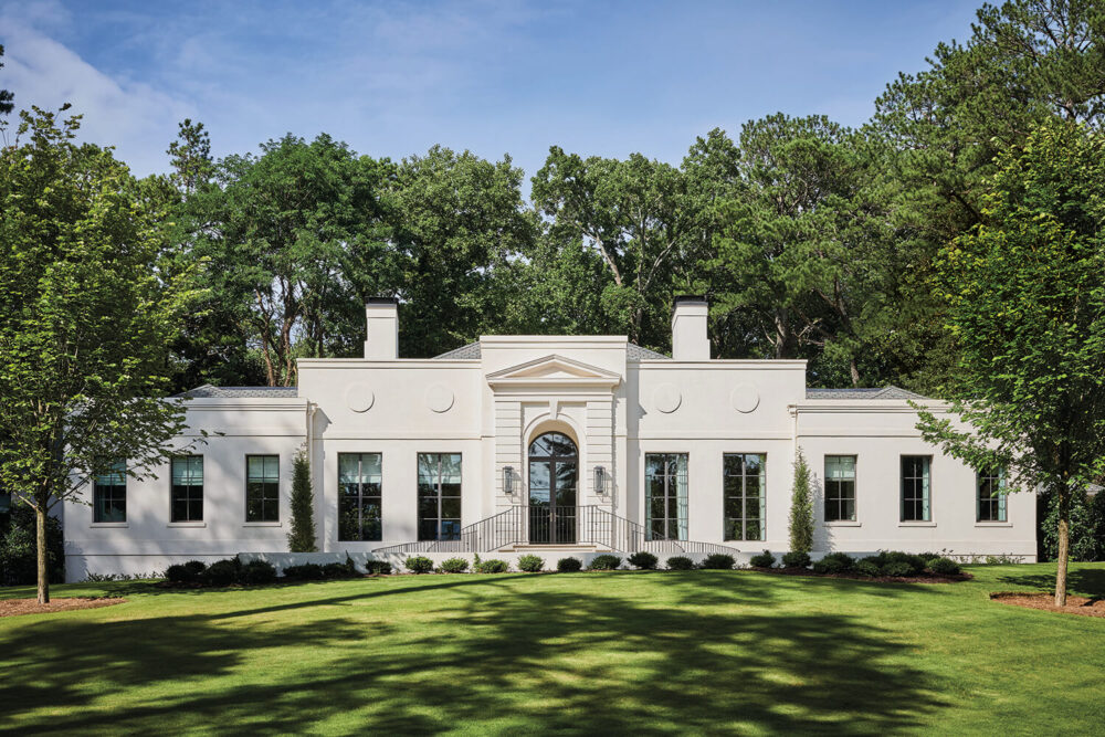 A white, single-story house with large windows and a central arched doorway, set in front of a manicured lawn and surrounded by tall trees.