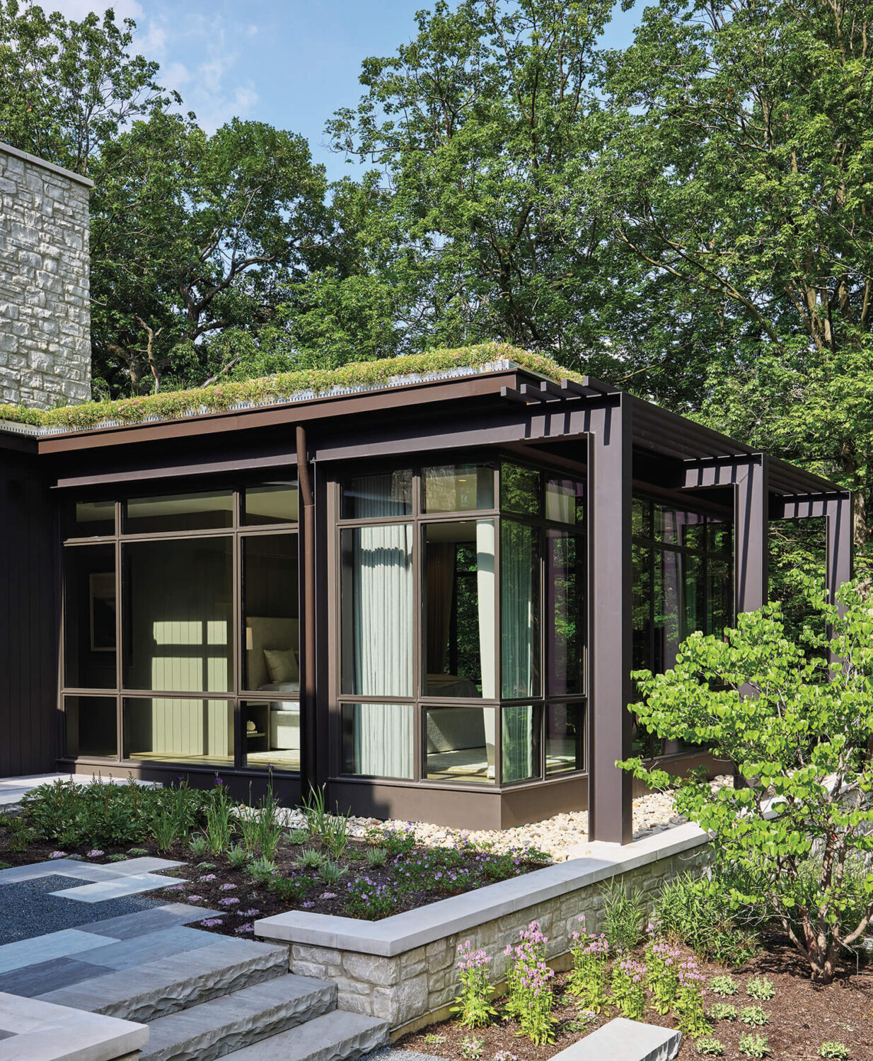 Modern house exterior with large glass windows, stone and metal accents, flat green roof, and landscaped garden surrounded by trees.