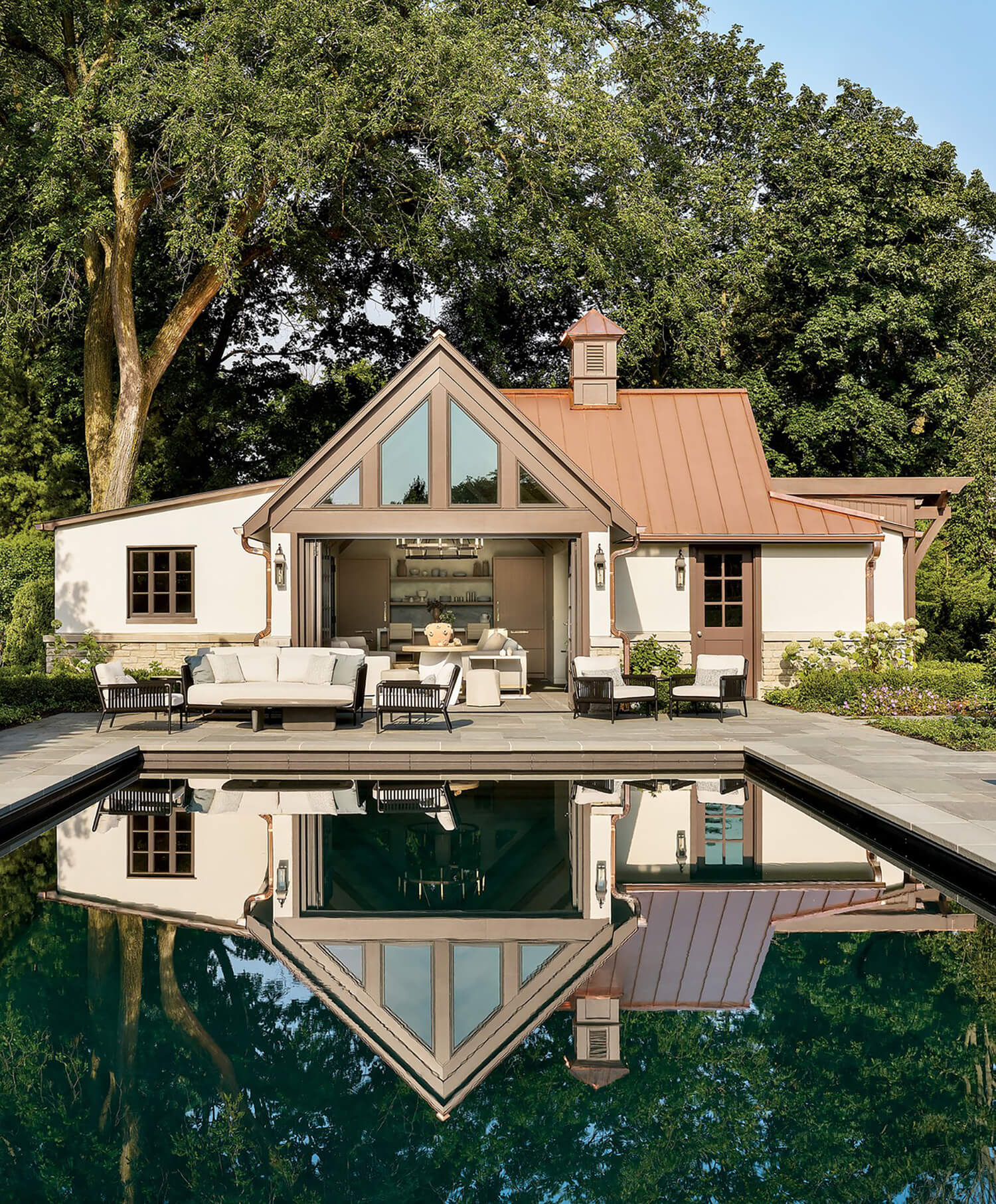 Small modern house with large windows and copper roof is reflected in a rectangular pool; patio furniture is arranged on the poolside terrace, surrounded by trees.