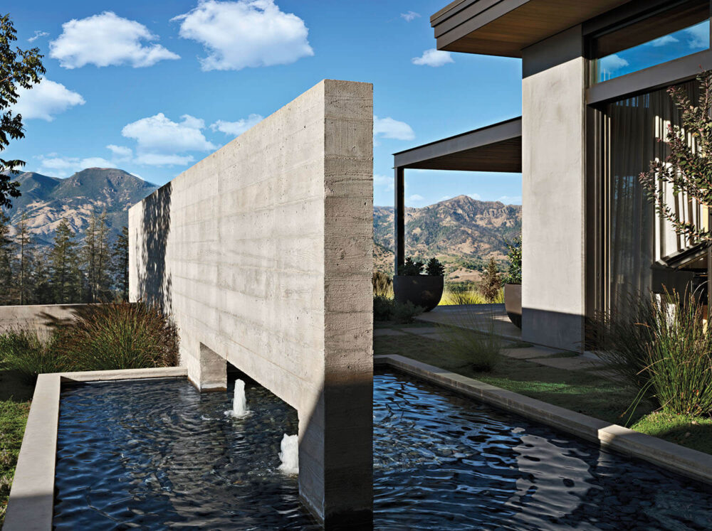 Modern concrete water feature with two fountains, set beside a contemporary building, with mountains and blue sky in the background.
