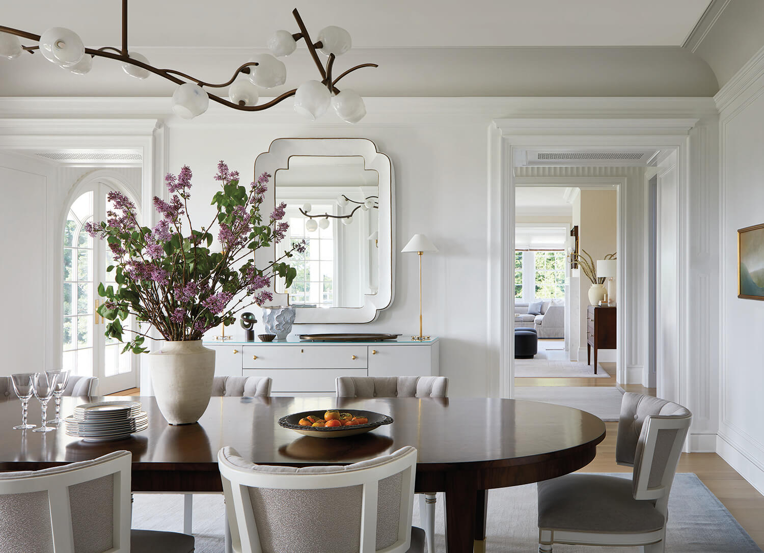 A dining room with a round wooden table, six upholstered chairs, a large vase of purple flowers, and a white sideboard with mirrors. A living room is visible through an open doorway.