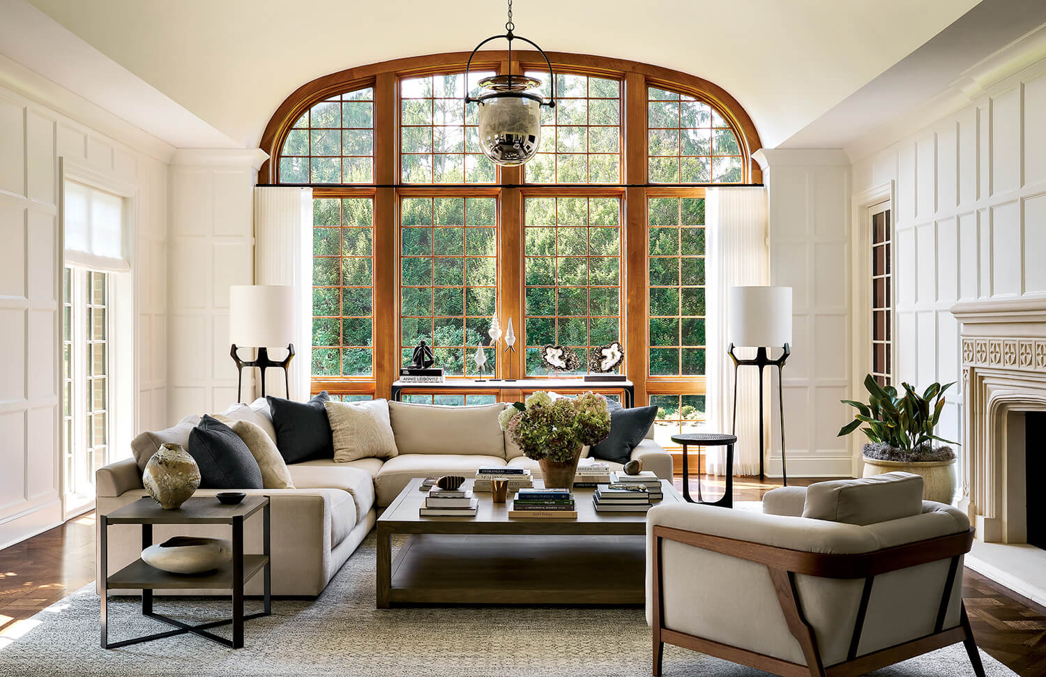 Elegant living room with large arched window, beige sofas, modern decor, two tall lamps, a coffee table with books and plants, and a fireplace on the right wall.
