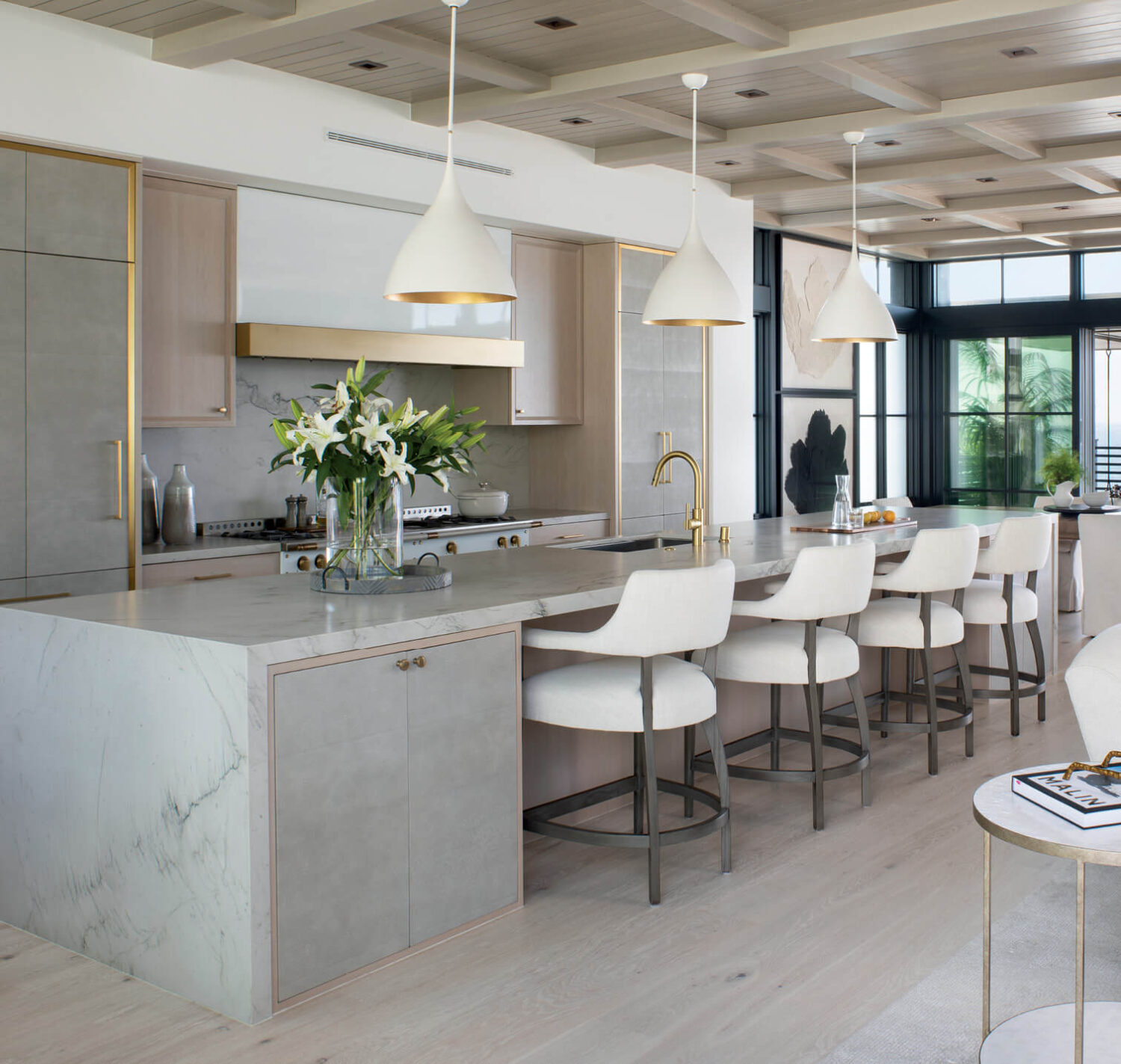 Modern kitchen with a marble island, four white barstools, pendant lights, stainless steel appliances, and a vase of white lilies on the counter. Light wood floors and large windows.