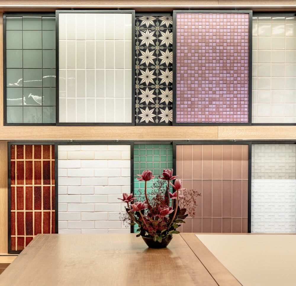 Display of various decorative wall tiles in different colors and patterns behind a wooden table with a small flower arrangement in the center.