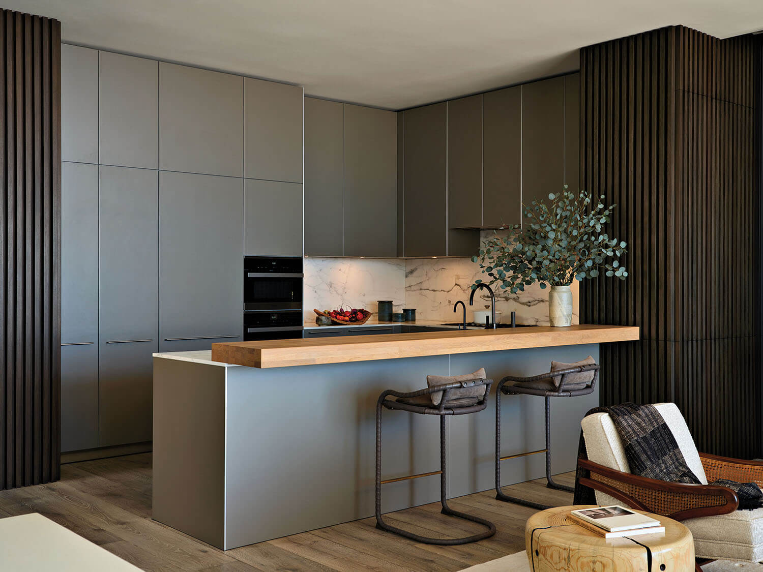 Modern kitchen with gray cabinets, wood-accented island, two bar stools, marble backsplash, black fixtures, and a vase with green branches on the counter.