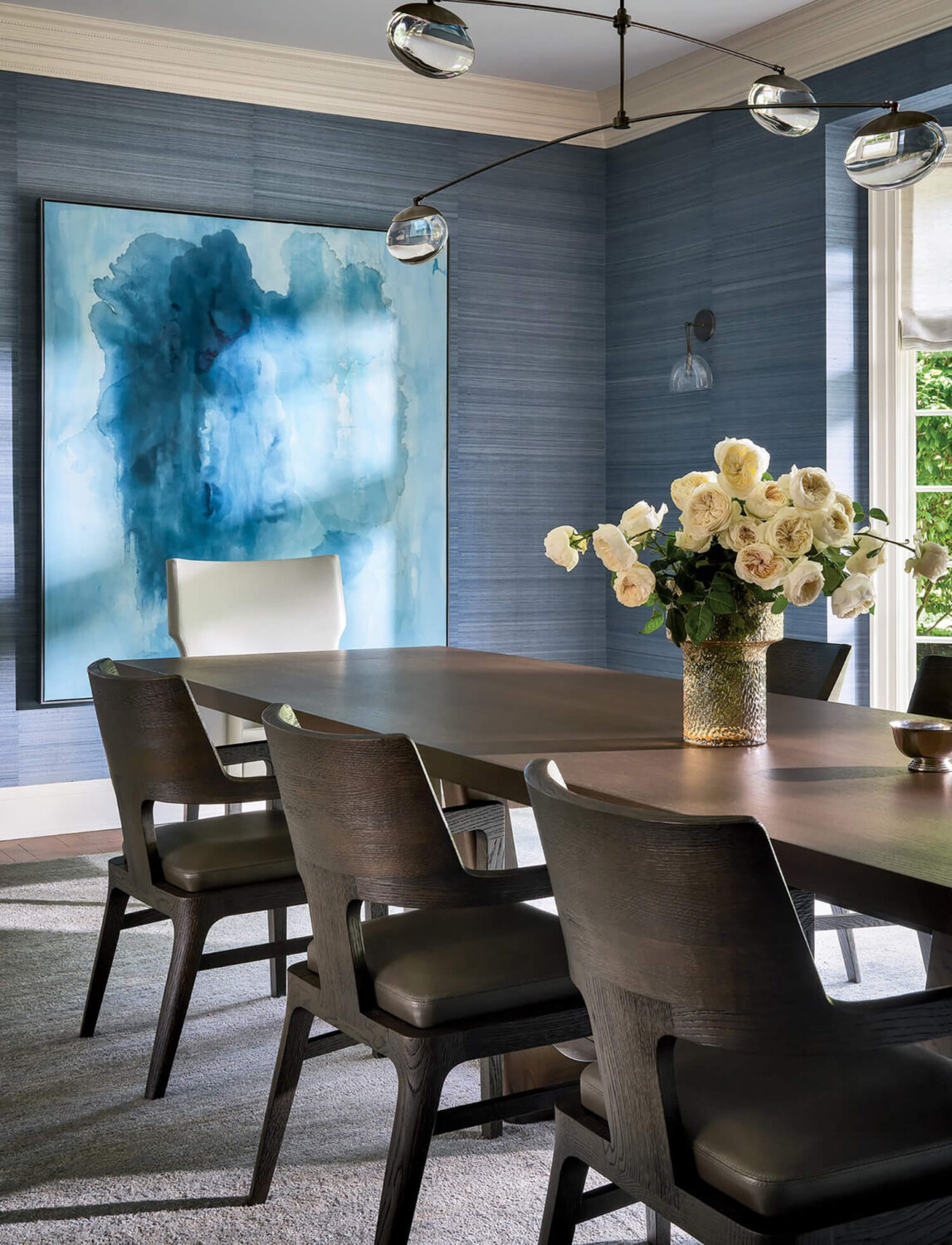 Modern dining room with dark wooden table and chairs, a vase of white roses, blue textured walls, large abstract blue art, and a contemporary light fixture.