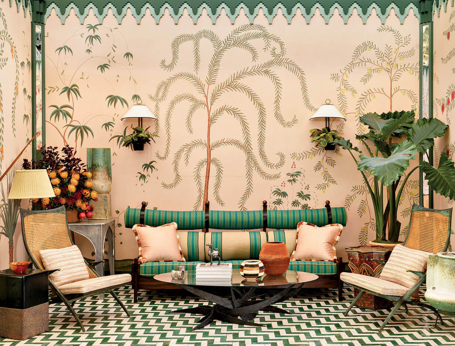 A tropical-themed room with palm murals, green-striped sofa, wicker chairs, zigzag floor, potted plants, wall sconces, and a black coffee table with books and vases.