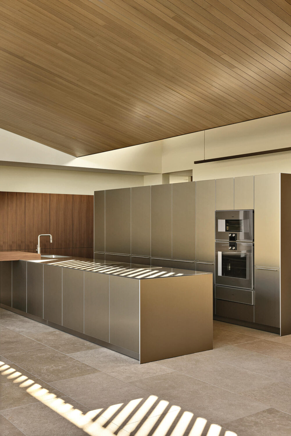Modern kitchen with metallic cabinets, built-in appliances, wood accent wall, minimalistic fixtures, and a wood-paneled sloped ceiling. Sunlight casts shadows on the island and floor.