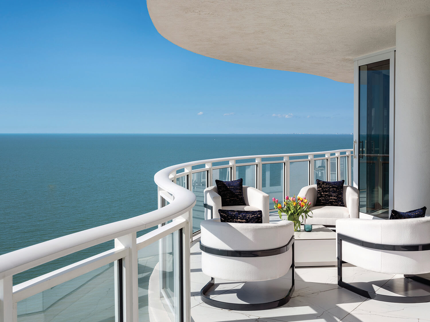 Modern balcony with white chairs and a table overlooking a calm ocean under a clear blue sky; glass railings and a vase of flowers on the table.