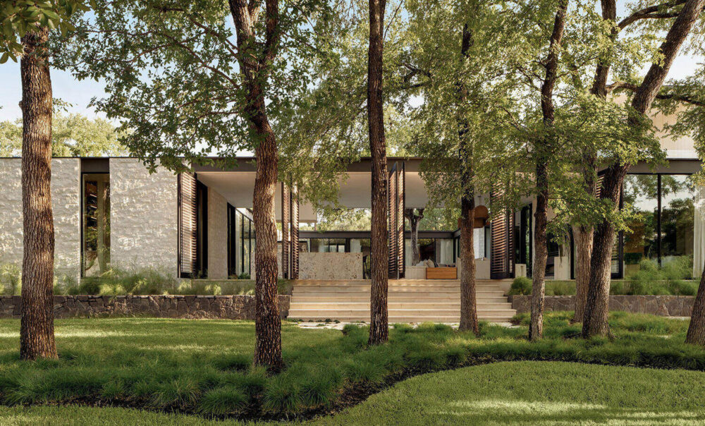 Modern house with large glass windows and stone walls, surrounded by green grass, tall trees, and landscaped plants in the foreground.