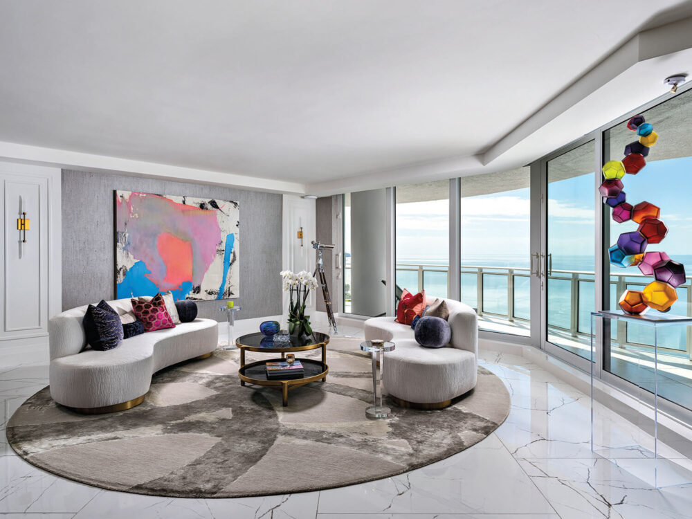 Modern living room with curved white sofas, abstract wall art, marble floor, round rug, and floor-to-ceiling windows overlooking the ocean. Colorful geometric sculpture by the window.