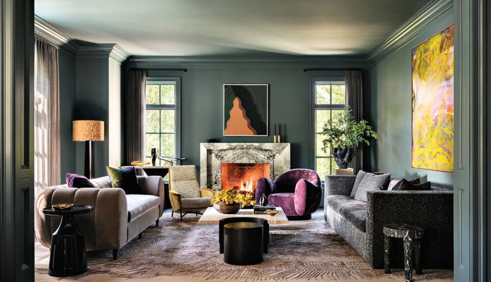 Modern living room with green walls, marble fireplace, abstract art, stylish furniture in neutral and purple tones, and large windows letting in natural light.