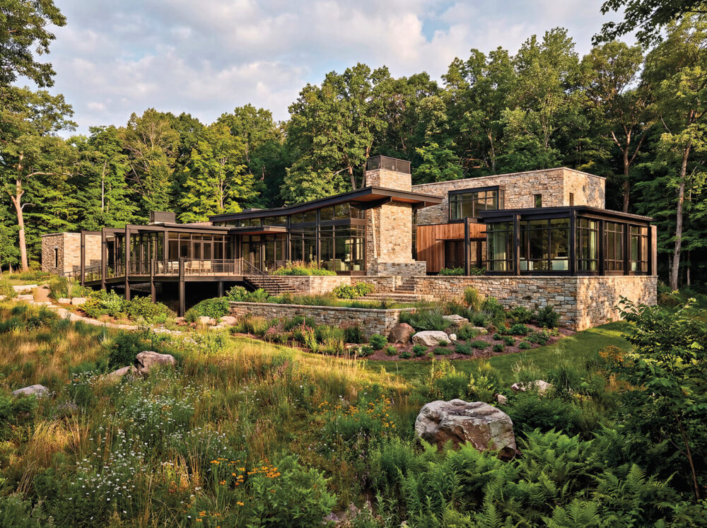 Modern stone and glass house with multiple angular sections, surrounded by dense trees and a landscaped garden with rocks and wildflowers.