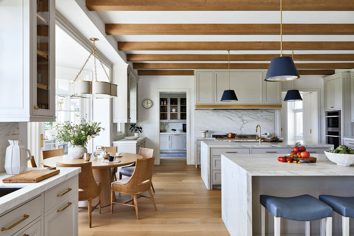 Modern kitchen with white cabinets, marble countertops, wood beams, a round dining table with wicker chairs, and two blue pendant lights above an island. Fruit and flowers are arranged on surfaces.