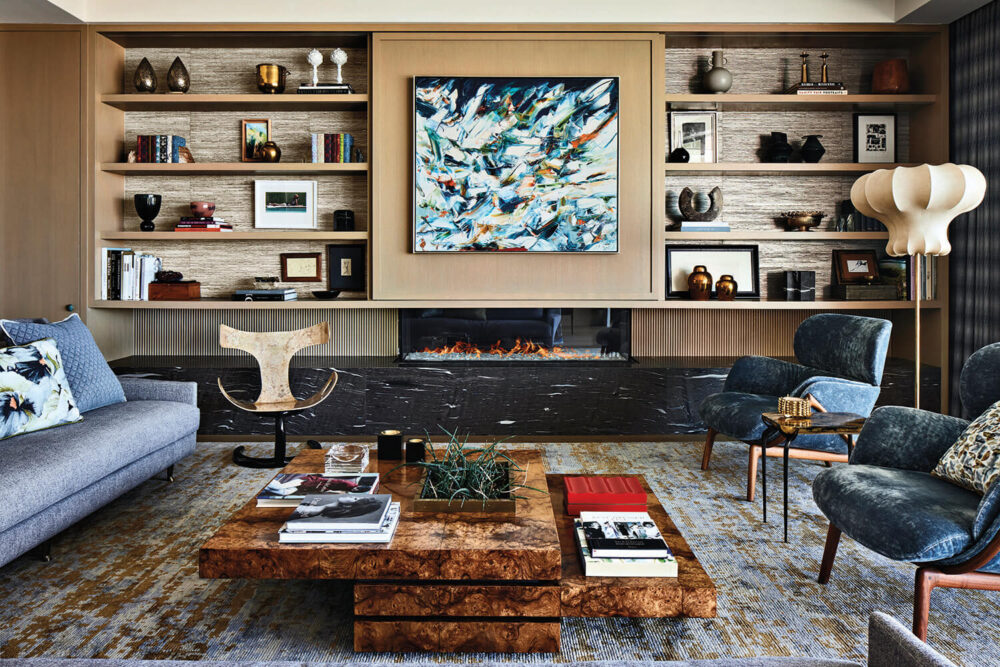 Modern living room with abstract art above a fireplace, built-in shelves with decor, a gray sofa, blue chairs, wooden coffee table with books, and a sculptural floor lamp.