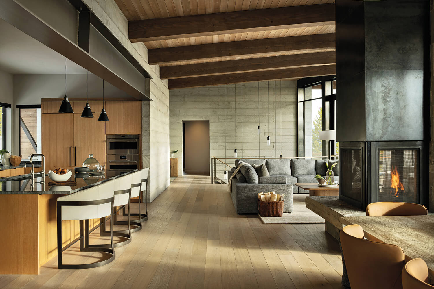 Modern open-concept living area with wood beams, kitchen island, gray sectional sofa, fireplace, and large windows letting in natural light.