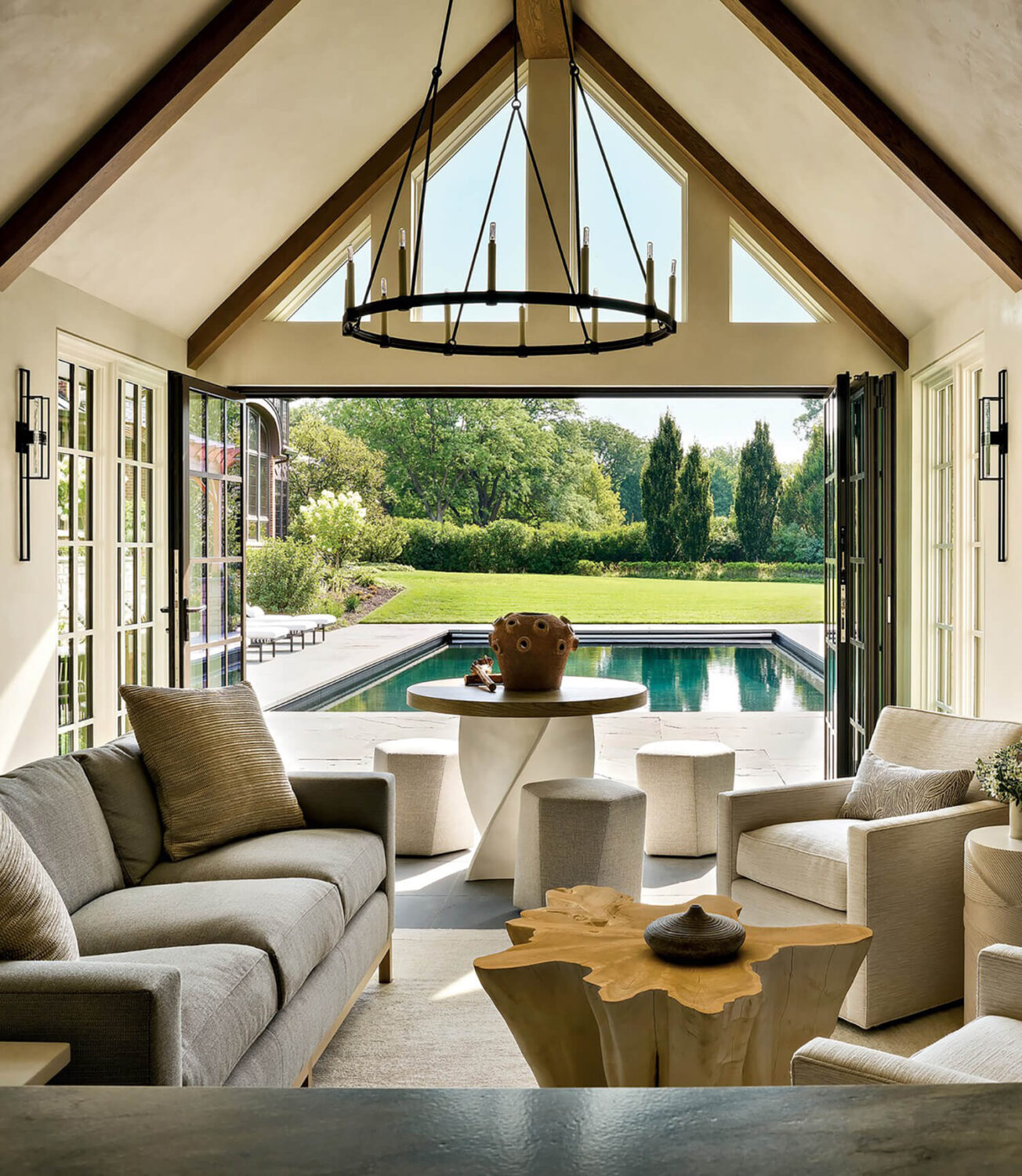 Bright sitting room with neutral furniture and wooden accents opens onto a patio and pool, with lush green lawn and trees visible through large glass doors.