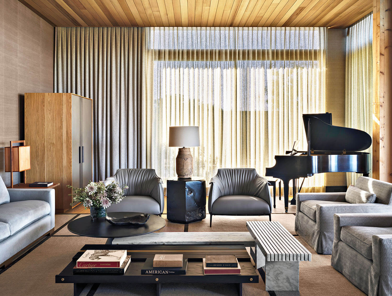 Modern living room with leather chairs, sofas, a round coffee table, books, lamp, cabinet, and a grand piano near floor-to-ceiling windows with sheer curtains and wooden ceiling.