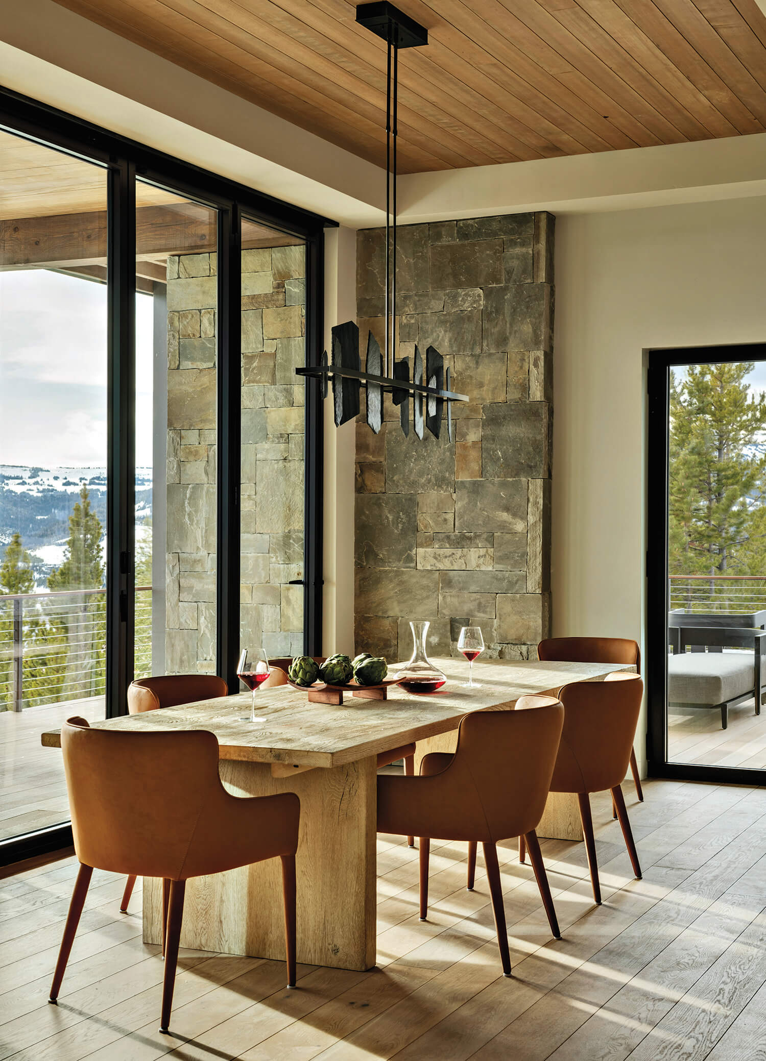 A modern dining room with a light wood table, six tan chairs, stone accent wall, large windows, and a black geometric chandelier.