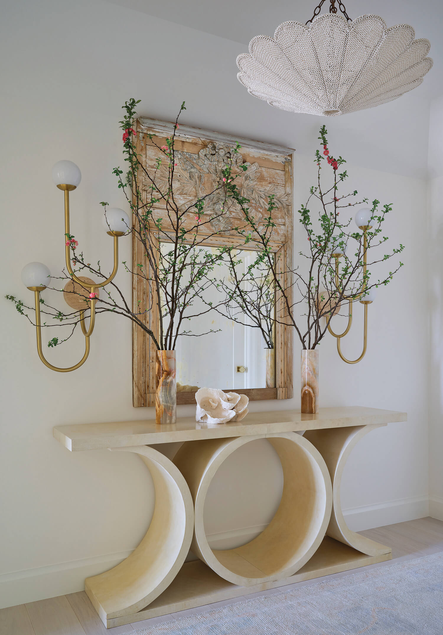 A modern entryway features a sculptural console table, two vases with flowering branches, a large mirror, gold wall sconces, and a white textured chandelier.