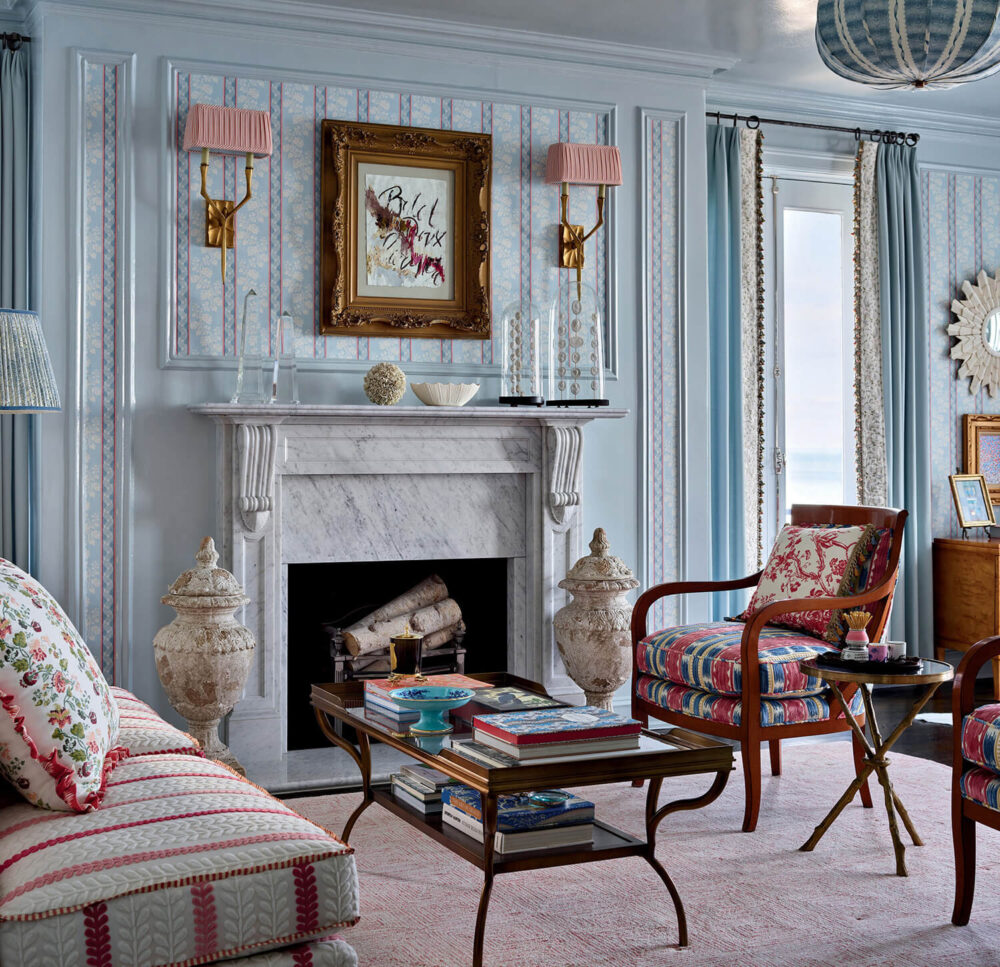 Elegant living room with a marble fireplace, armchairs, a glass coffee table with books, patterned sofa, framed art, and pastel decor. Large windows bring in natural light.