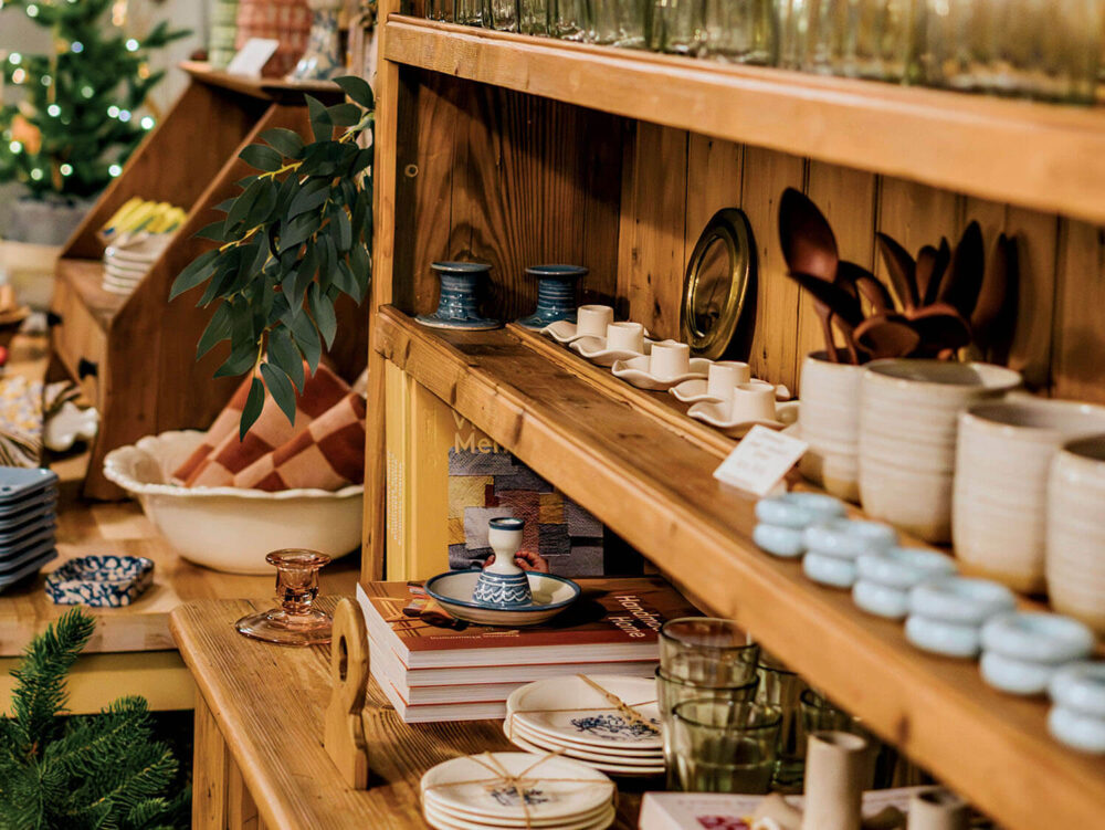 Wooden shelves display various ceramics, glassware, and dishes in a store. Stacked plates, cups, and decorative items are neatly arranged. A plant and holiday lights are visible in the background.