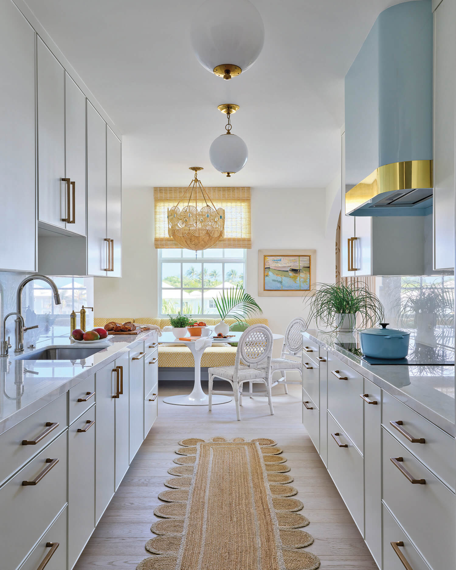 Modern galley kitchen with white cabinets, brass handles, a woven rug, and a breakfast nook by a window with natural light. A blue range hood and plants add color accents.