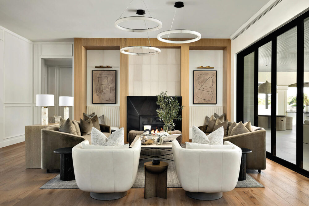 Modern living room with two white chairs, four brown sofas, a central black table, abstract wall art, and circular pendant lights above a fireplace. Large windows line the right side.
