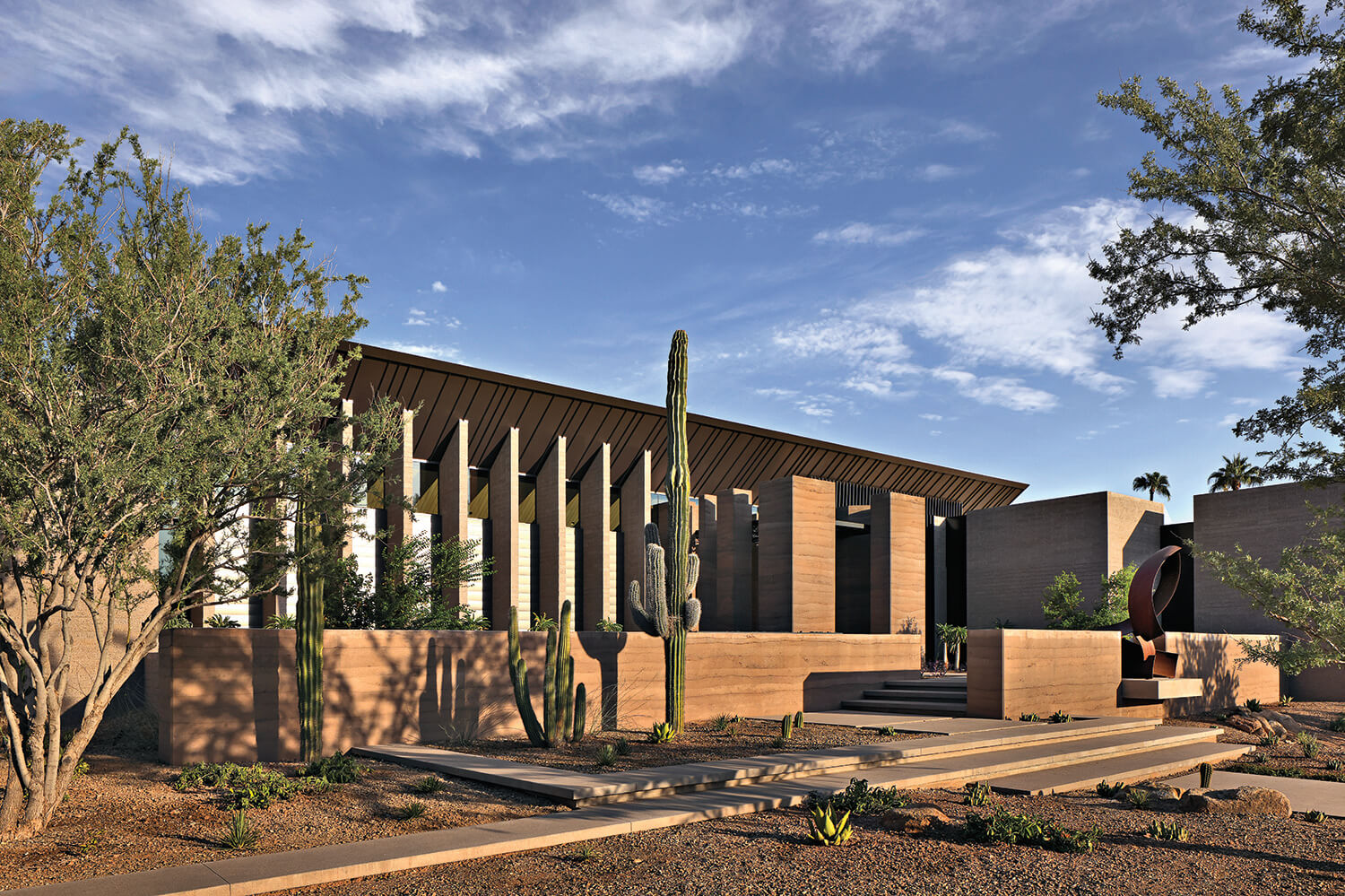 Modern building with tall vertical columns, desert landscaping, cacti, and clear blue sky in the background.