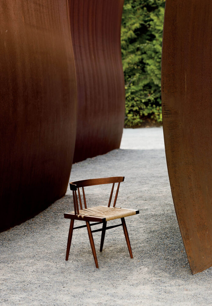 A wooden chair with a woven seat is placed on gravel between large rust-colored, curved metal walls with greenery in the background.