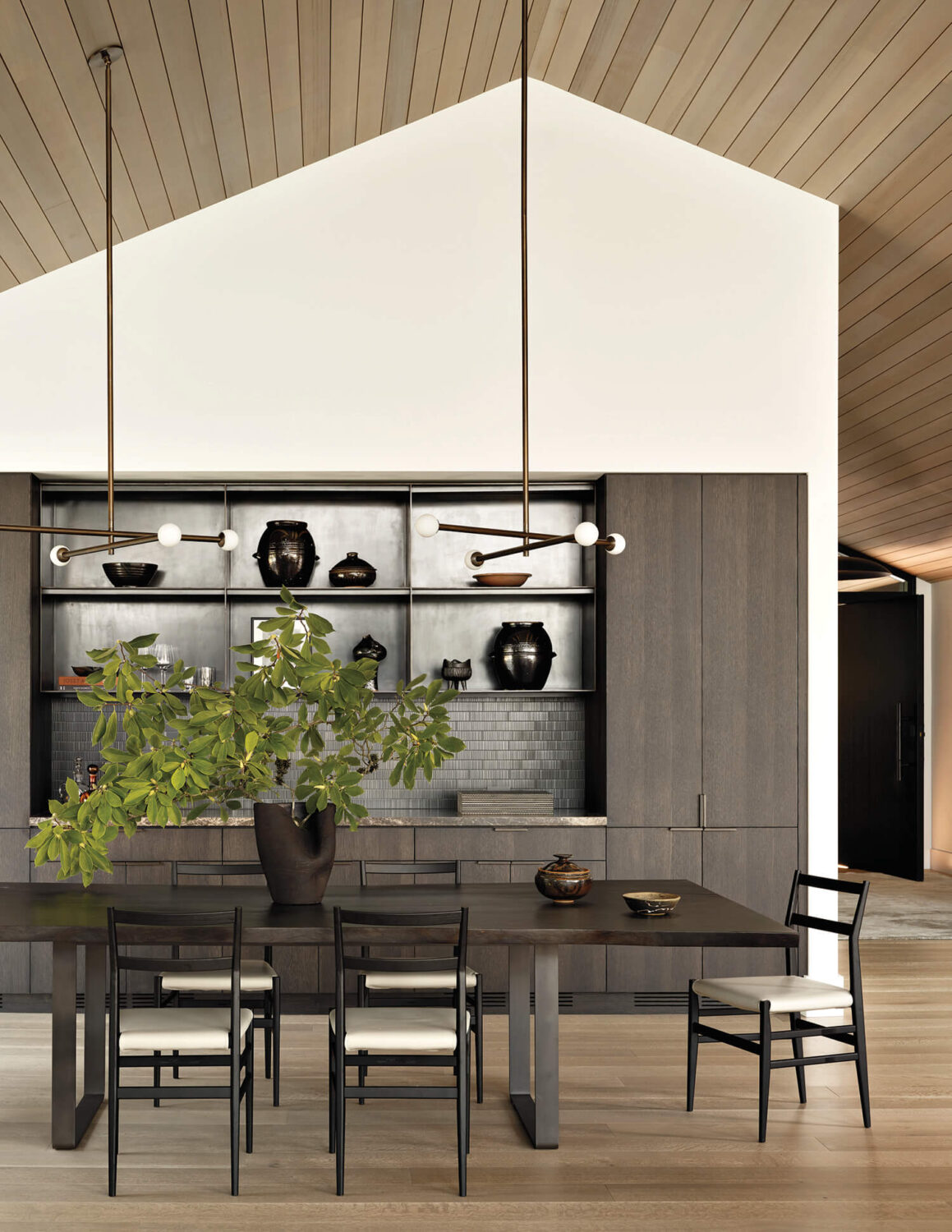 Modern dining room with a dark wooden table, six chairs, a large vase with green branches, and shelves displaying black pottery against a light wood ceiling and floor.