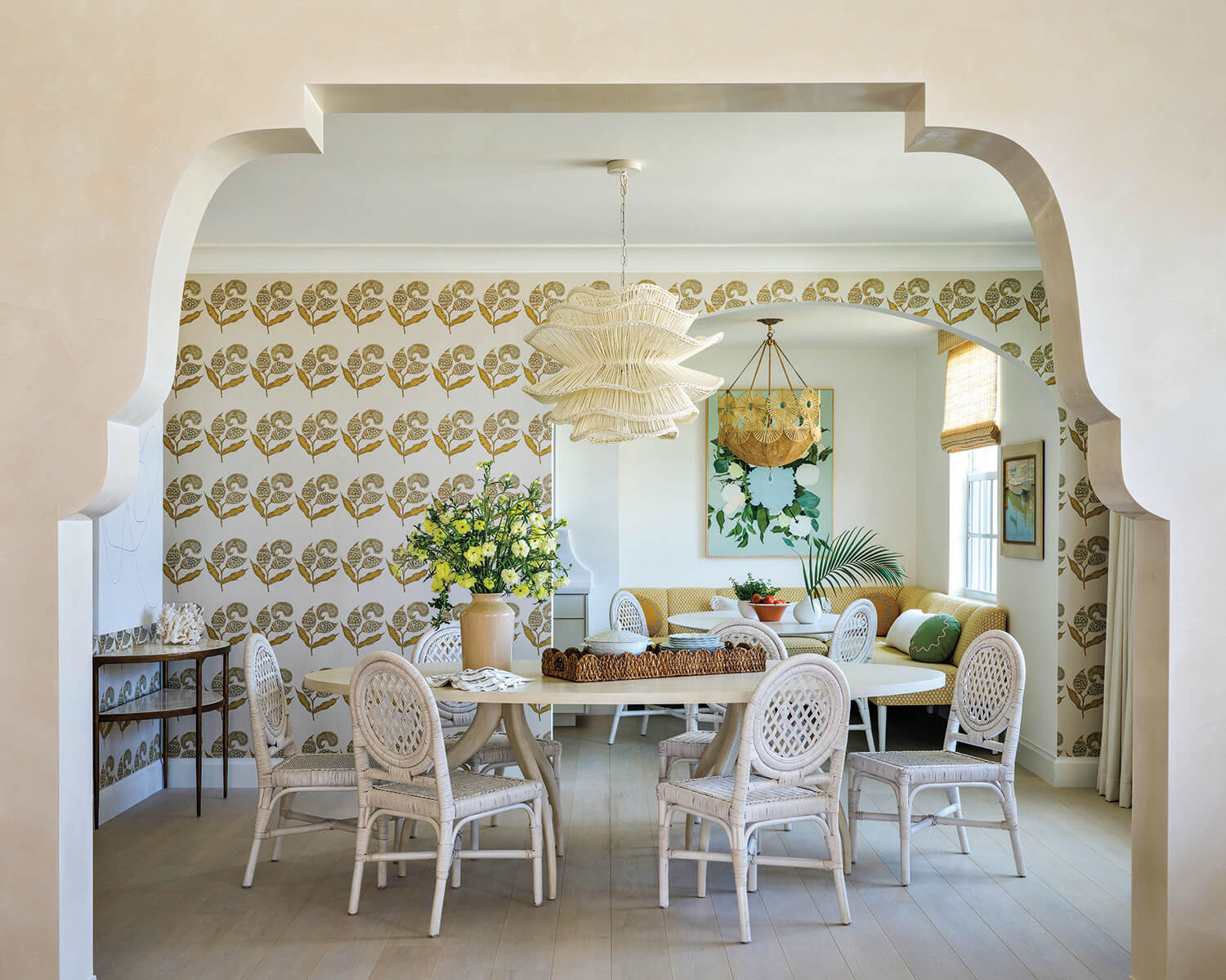 Dining room with white wicker chairs, oval table, patterned wallpaper, floral arrangement, pendant light, and archway; window seating with pillows and wall art in background.