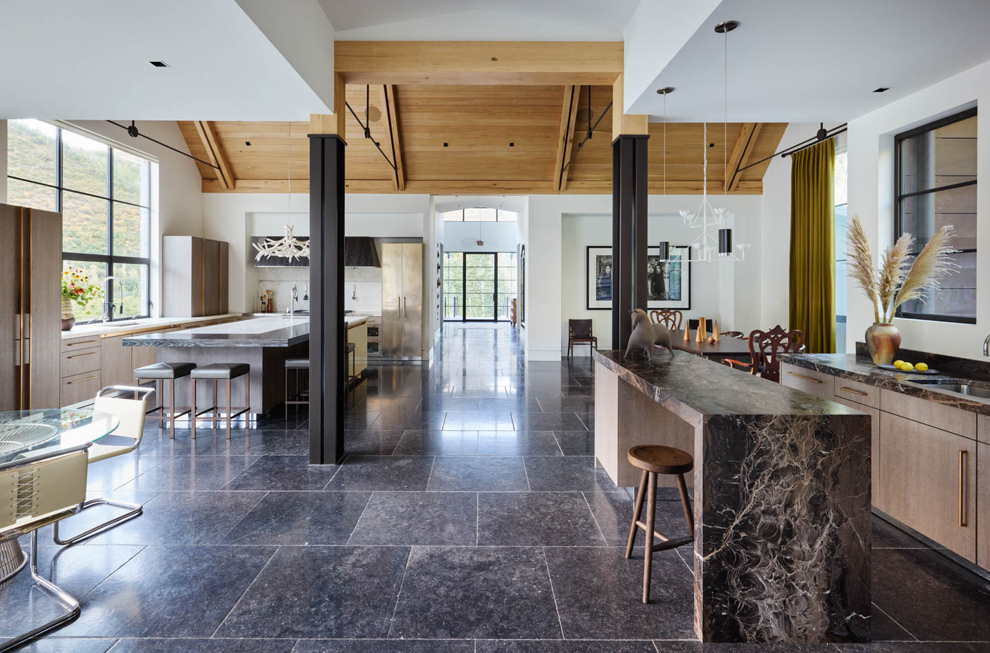Modern kitchen with high wood ceilings, marble countertops, large windows, dark tile flooring, and a mix of dining and bar seating, featuring neutral and natural tones.
