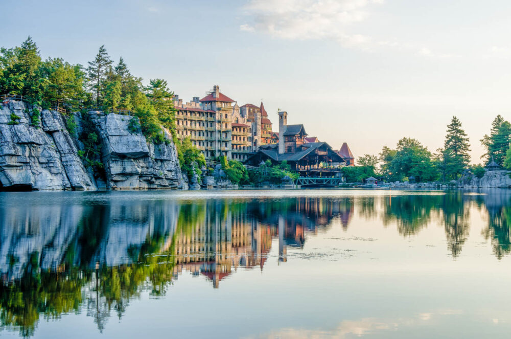 Large stone building with towers sits beside a calm lake, surrounded by trees and rocky cliffs, with clear reflections in the water under a blue sky.