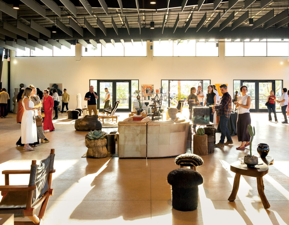 People gather in a spacious, sunlit room with modern furniture and decor displays, engaging in conversation and browsing various items.
