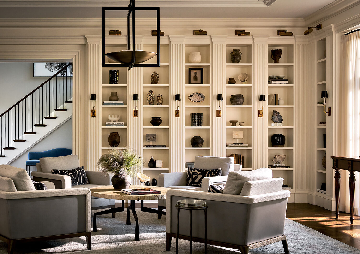 Elegant living room with four gray armchairs around a round table, built-in shelves filled with decor, and a staircase visible in the background.