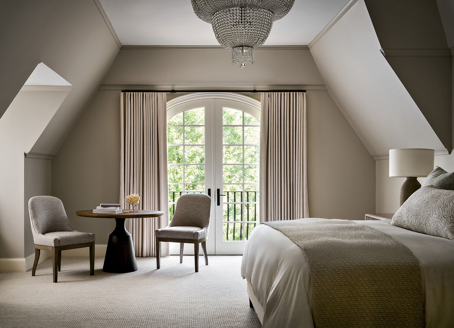 A neutral-toned bedroom with a bed, two chairs at a round table, a large arched window with curtains, and a chandelier.