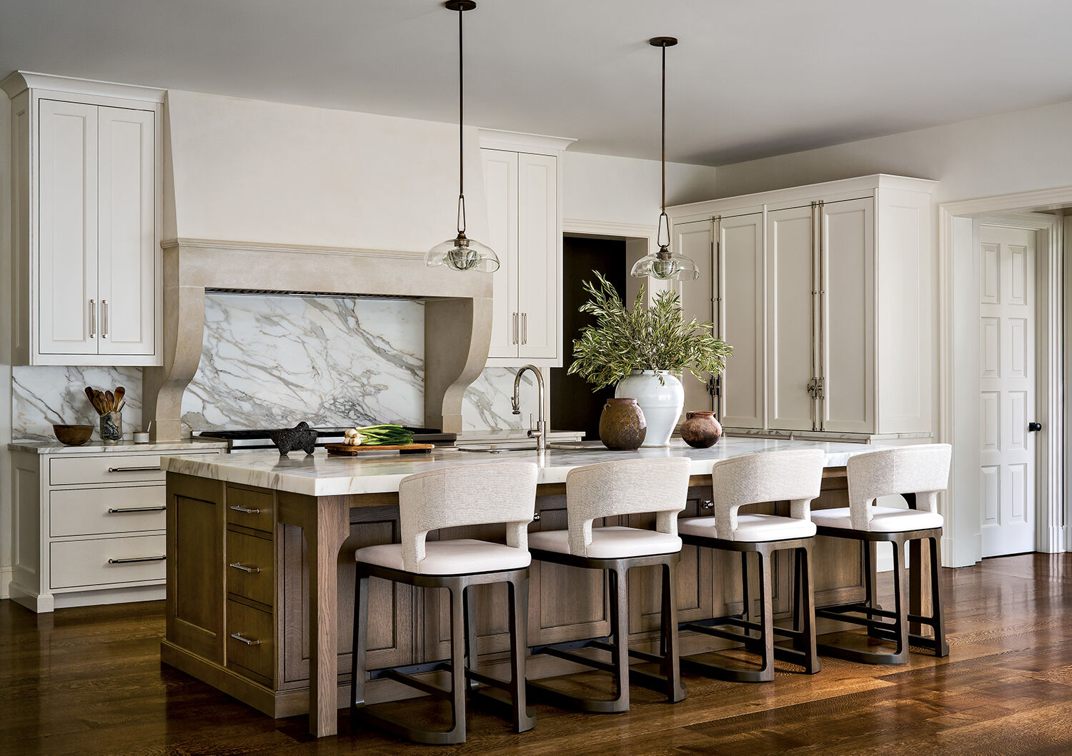 Modern kitchen with a large island, four upholstered barstools, marble backsplash, pendant lights, white cabinets, wood floors, and decorative vases on the counter.