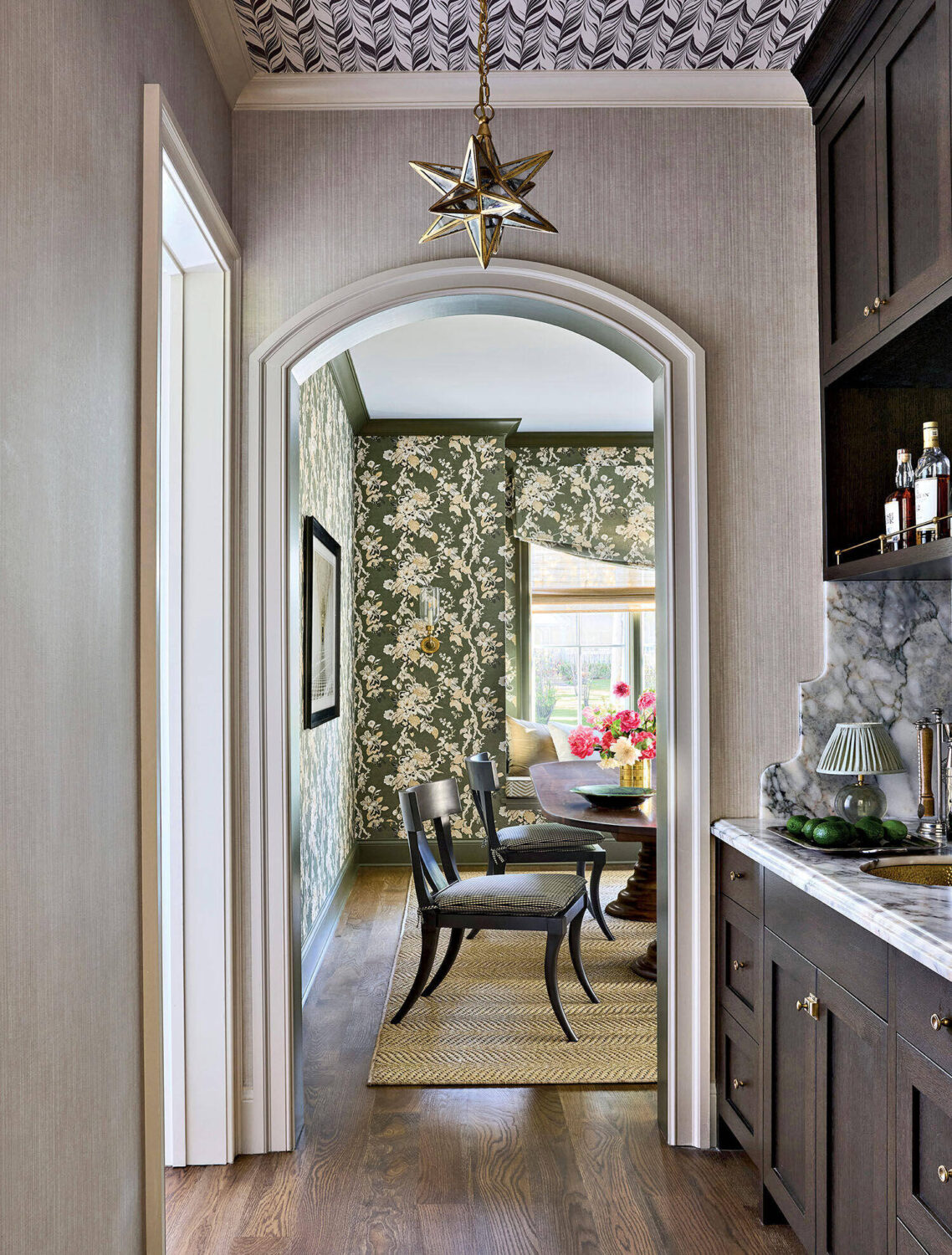 View through an arched doorway into a dining room with floral wallpaper, black chairs, a table with flowers, and natural light from a window.