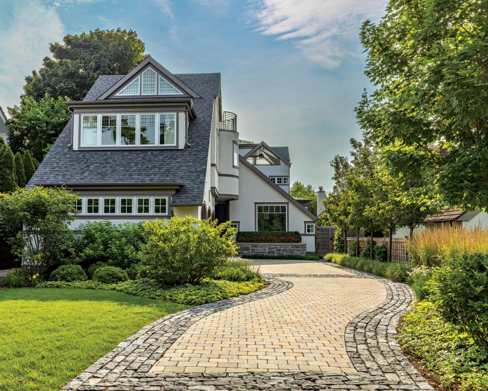 Modern two-story house with large windows, a landscaped garden, and a curved stone driveway, surrounded by green trees and a sunny sky.