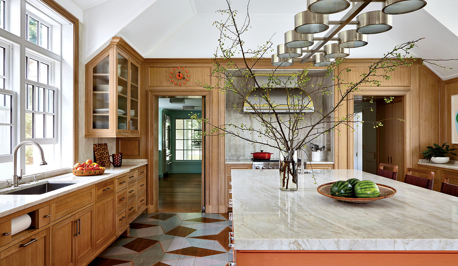 Spacious kitchen with wooden cabinets, marble island, modern lighting, a large vase with branches, and watermelons on the counter. Sunlight streams in through wide windows.