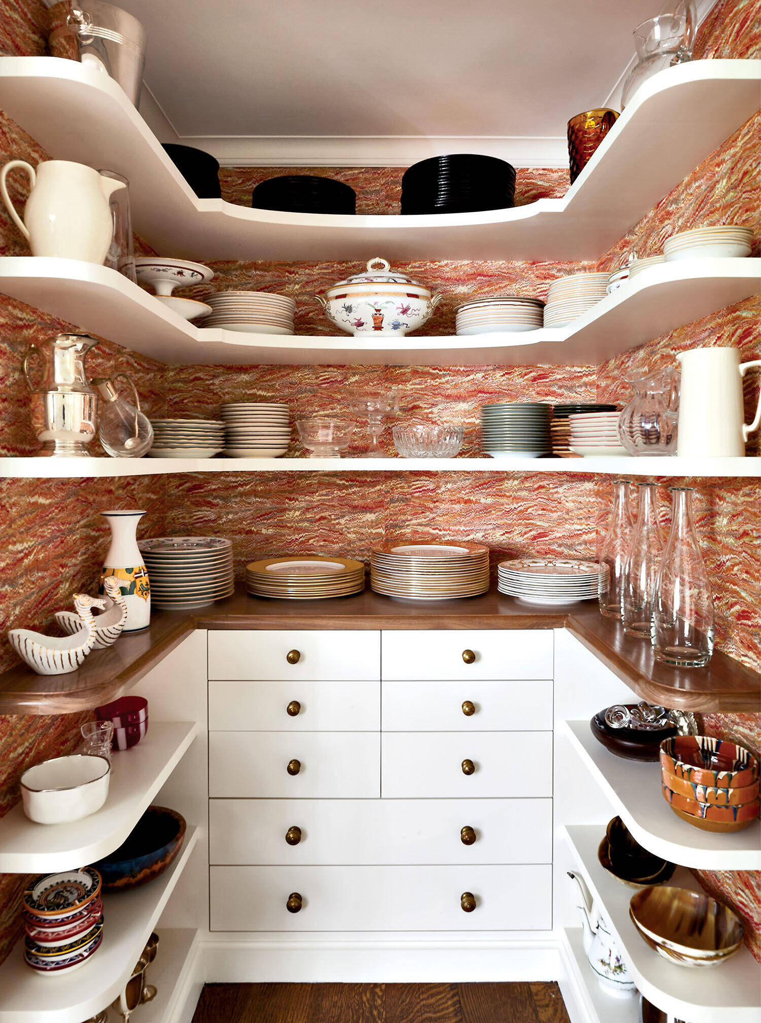 A neatly organized pantry with white shelves holding various plates, bowls, glasses, and pitchers, set against a colorful, marbled wallpaper background.